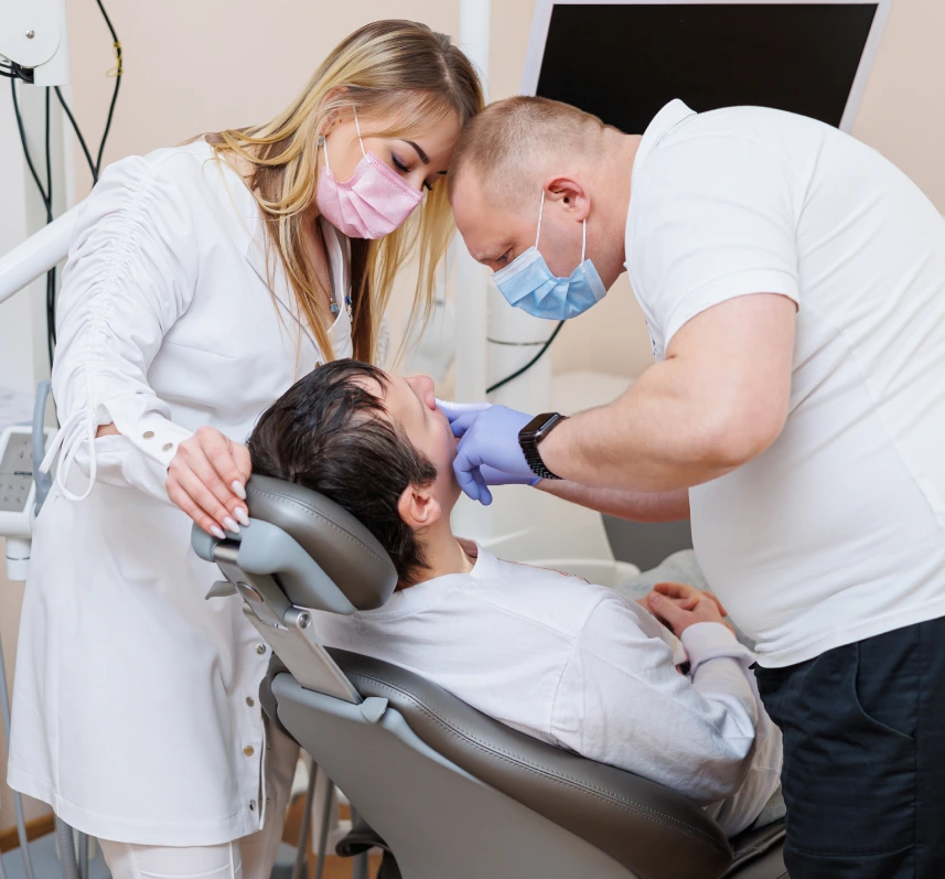 Clinician examining a patient's mouth while an assistant stands nearby in a dental clinic