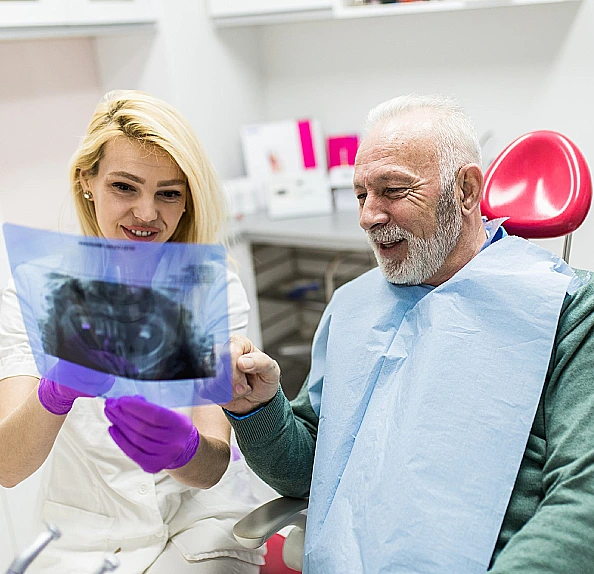 Clinician holding a dental X-ray film while an older man in a treatment chair points to it