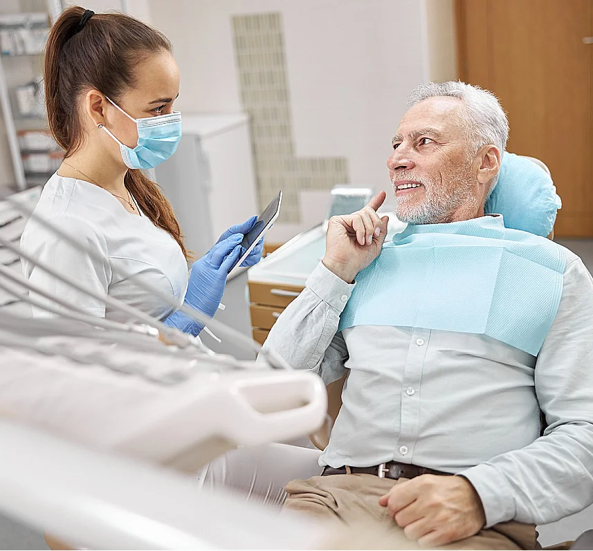 Clinician in a surgical mask holding a tablet while speaking with a man in a dental chair pointing to his cheek