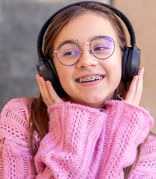 Smiling girl wearing round glasses, over-ear headphones, and dental braces in a pink sweaterphones dental braces