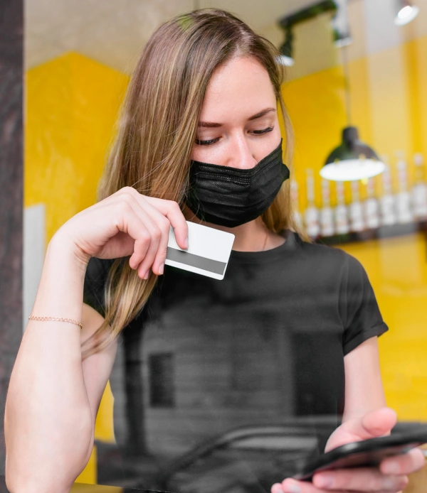 Woman wearing a black face mask holding a blank plastic card and using a smartphone