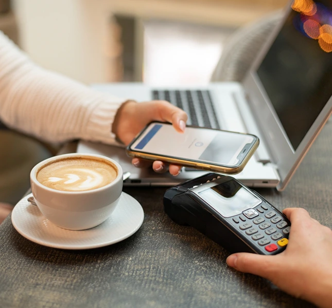 Person holding a smartphone over a payment terminal beside a laptop and coffee